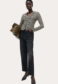 Donna che indossa una blusa a righe con volant, jeans neri a gamba dritta, tacchi a punta neri, mentre tiene una grande borsa a tracolla in pelle marrone.