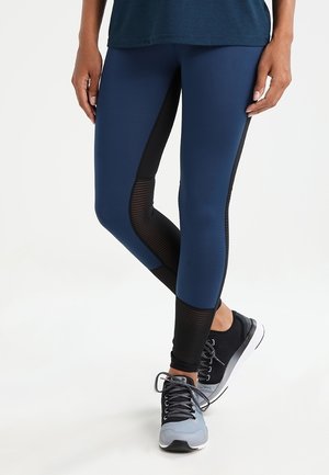 Navy blue leggings dengan sisipan jala hitam dan tekstur halus; dipadukan dengan sepatu olahraga hitam dan abu-abu.