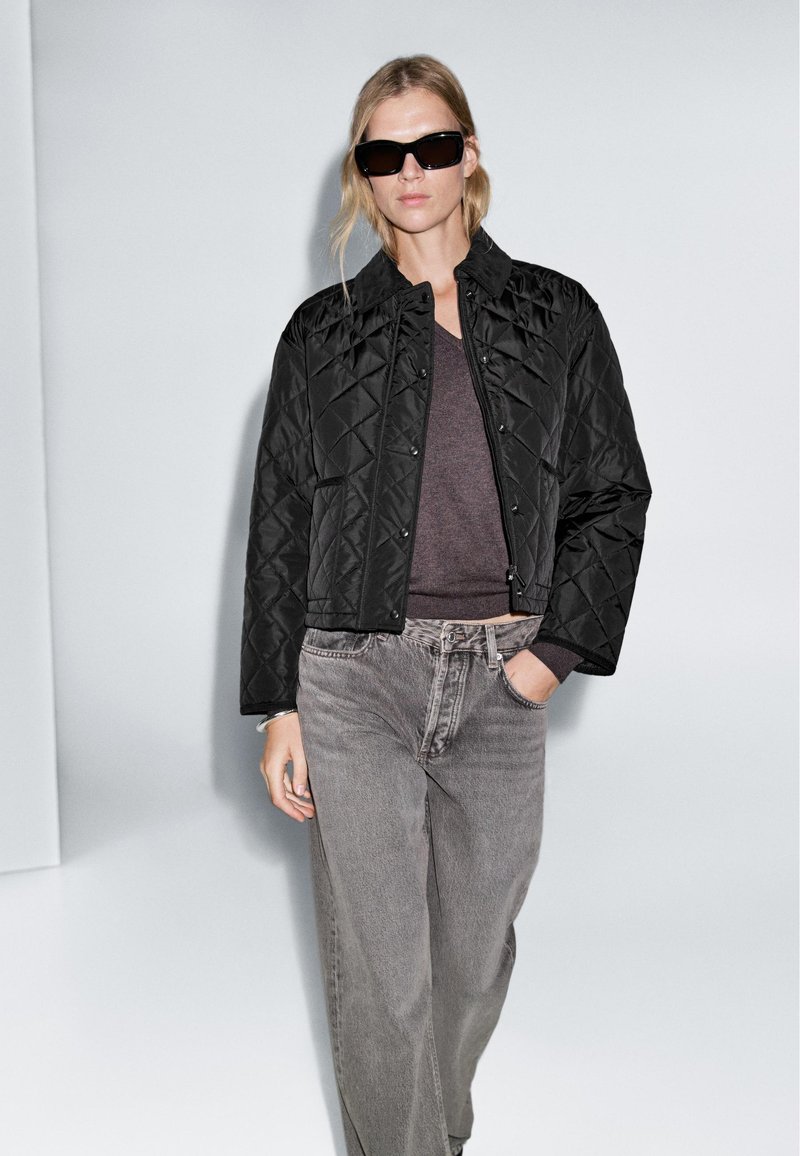 Veste matelassée noire avec boutons-pression, associée à un pull gris foncé et à un jean taille haute gris clair. Mannequin portant des lunettes de soleil noires.