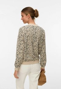 Sieviete ar matiem sasietiem bulciņā valkā bēša un melnas krāsas rakstainu džemperi, baltas bikses, zelta auskarus un tur rokās brūnu zamša somiņu.