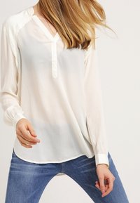 Žena v dlouhém rukávu průhledné bílé blůze s výstřihem do V a modrých džínách, pózující na jednobarevném světlém pozadí.