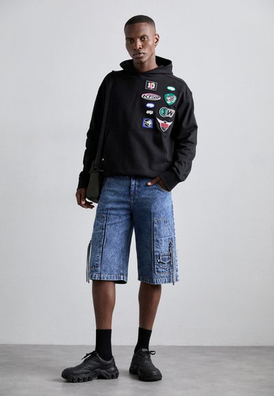 Black hoodie with various patches, paired with blue denim cargo shorts featuring side zippers, worn with black sneakers and crew socks.