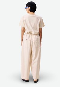 Persona vistiendo pantalones sueltos de color beige claro con bolsillos traseros, camisa de manga corta beige a juego, sandalias negras y gafas de sol azules, de espaldas.