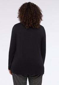 Osoba s kudrnatými vlasy, oblečená v černém tričku s dlouhým rukávem a šedých texturovaných kalhotách, stojí zády k prázdnému světlému pozadí.