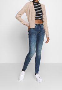 Krémový pletený kardigan s knoflíkovým zapínáním, nošený přes černobílé pruhované crop top, v kombinaci s modrými skinny džínami a bílými teniskami.
