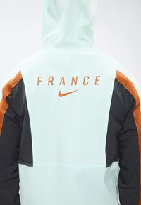 Οπίσθια όψη ενός μπουφάν με τη λέξη "FRANCE" και το πορτοκαλί σήμα Nike, που διαθέτει μπλοκ χρωμάτων σε ανοιχτό μπλε, μαύρο και πορτοκαλί στα μανίκια.