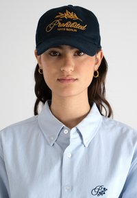 Laivastonsininen lippalakki, jossa on keltainen "Kielletty" -kirjonta ja lehdesuunnittelu. Vaaleansininen napillinen paita, jossa on huomaamaton logo-ystävällinen yksityiskohta rinnassa.