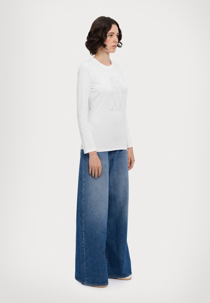 Top bianco in cotone a maniche lunghe con stampa grafica al centro, abbinato a jeans in denim blu a gamba larga con una lavatura sfumata.