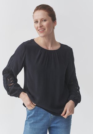 Zwarte blouse met ronde halslijn, pofmouwen met kantdetails. Zacht materiaal met een losse pasvorm. Draagt met lichtblauwe jeans.
