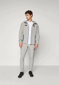 Világosszürke sportdzseki fekete részletekkel és Adidas logóval, hozzá illő világosszürke nadrággal, fekete sportcipővel viselve.