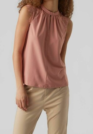 Femme portant un haut sans manches rose poussiéreux avec des épaules en dentelle et un pantalon beige sur un fond gris uni.