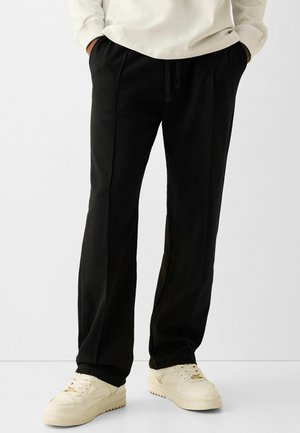 Partie inférieure du corps portant un pantalon noir à cordon et des baskets blanc cassé, les mains dans les poches, debout contre un fond clair uni.