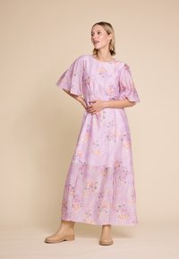 Lilla maxikjole med flagrende ærmer, prydet med et blomsterdesign i pink og grøn. Glat tekstur med en indsnævret talje, parret med beige sko.