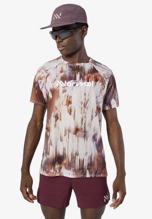 Hombre con gorra marrón, gafas de sol deportivas negras, camiseta de manga corta con estampado abstracto y pantalones cortos deportivos marrones, de pie con los brazos relajados.