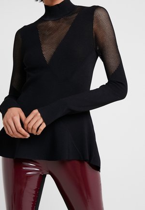 Femme portant un haut noir à manches longues en maille filet et tricot uni avec col montant, associé à un pantalon en vinyle bordeaux brillant.