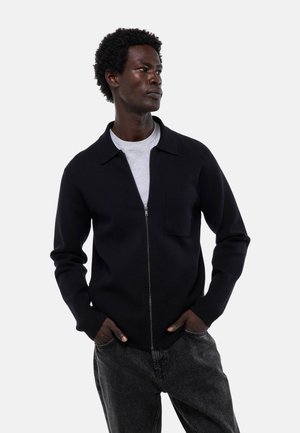 Pull noir à fermeture éclair avec col à revers, doté d'une poche poitrine et de poignets côtelés, porté par-dessus un t-shirt gris, associé à un jean foncé.