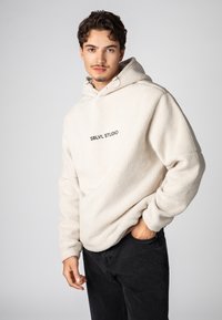 Crèmekleurige fleece hoodie met een frontale kangoeroezak en geprinte "SBLVL STUDIO" logo. Gecombineerd met zwarte broek, eenvoudig design.