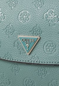 Tyrkysová texturovaná kabelka s reliéfními vzory a trojúhelníkovou stříbrnou destičkou s výrazným nápisem "GUESS USA".