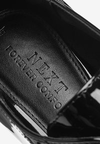 Intérieur de chaussure en cuir verni noir avec la marque embossée "NEXT FOREVER COMFORT" sur une surface texturée. Design élégant avec des bords lisses.