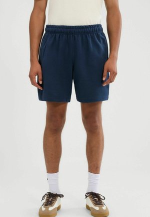 Mężczyzna ubrany w granatowe spodenki z elastycznym pasem, białe skarpetki oraz beżowe sneakersy z brązowymi sznurówkami, stojący na jednolitym jasnym tle.