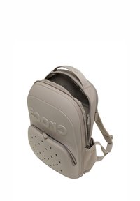 Mochila beige de Crocs con bolsillo frontal con cremallera y diseño perforado, compartimento principal parcialmente abierto que muestra el forro interior.