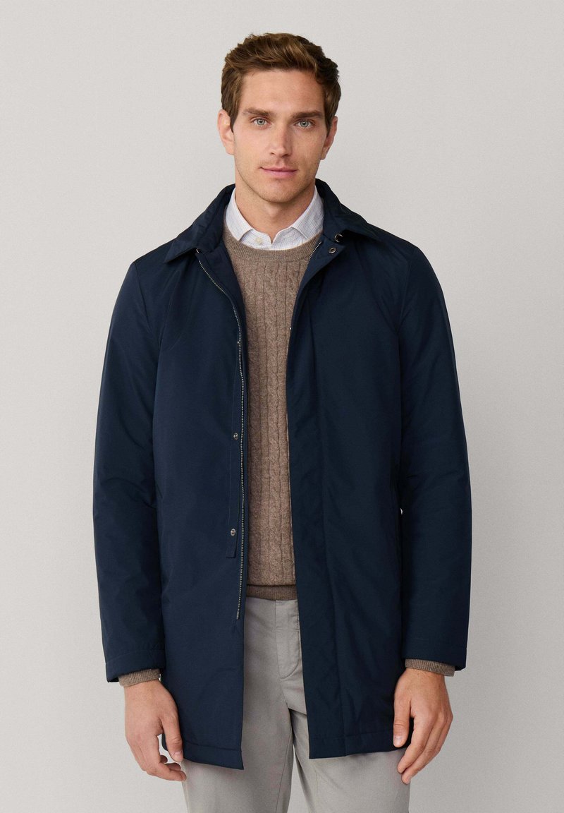 Marineblauer wasserdichter Mantel mit Kragen, Frontreißverschluss und Druckknöpfen. Über einem braunen Strickpullover und hellgrauen Hosen getragen.