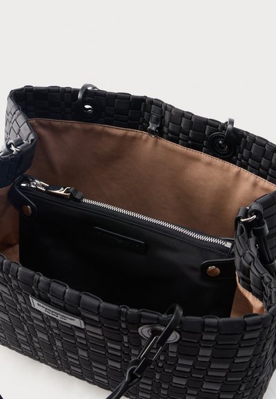 Μαύρη υφαντή τσάντα tote με καφέ εσωτερική επένδυση και αποσπώμενη μαύρη τσαντούλα με φερμουάρ στο εσωτερικό, παρουσιάζεται σε απλό φόντο.