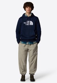 Sudadera navy con logo blanco, bolsillo tipo canguro; pantalones beige claros con puños elásticos; botas de senderismo negras y amarillas.