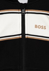 Haut à capuche zippé noir avec des rayures horizontales blanches et cuivrées. Présente un logo "BOSS" en marron clair sur le côté supérieur gauche.
