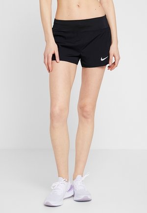 Femme portant un short de sport noir Nike et des chaussures de course blanches, debout devant un fond clair uni.