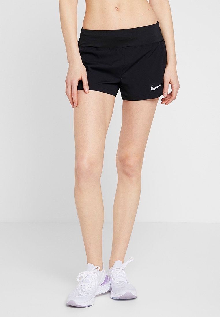 Femme portant un short de sport noir Nike et des chaussures de course blanches, debout devant un fond clair uni.