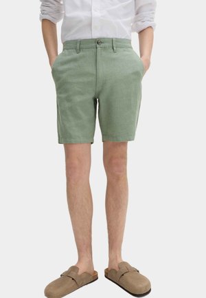 Hombre vistiendo pantalones cortos sastre de color verde claro, camisa blanca abotonada con las mangas remangadas y zapatos marrones sin cordones, de pie con las manos en los bolsillos.