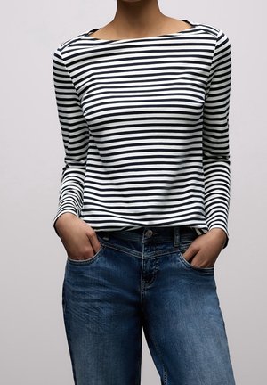 Person iført en langærmet sort og hvid vandretstribet skjorte stukket ned i blå jeans med hænder i lommerne mod ensfarvet baggrund.