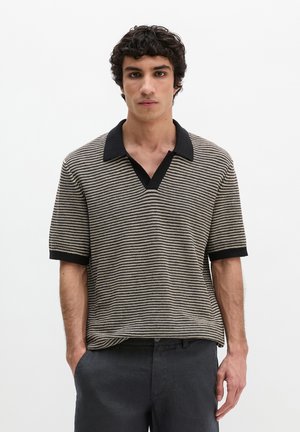 Jeune homme aux cheveux bouclés et foncés, portant un polo à manches courtes rayé noir et beige et un pantalon sombre, debout sur un fond blanc.