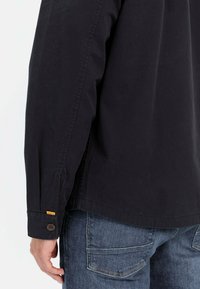 Zwarte katoenen shirt met een knoopmanchet en gele accenten; heeft een rechte snit en platte naden, gecombineerd met blauwe spijkerbroeken.