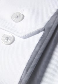 Camicia bianca con un tessuto texturizzato, con bottoni visibili e cuciture ordinate lungo i bordi, piegata con cura.
