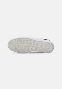 Talpa de sneaker albă cu un model texturat pentru aderență, având o suprafață netedă și o ușoară curbură spre vârf. Accente negre la călcâi.