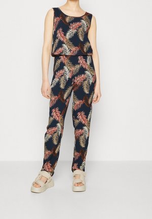 Frau trägt dunkelblauen ärmellosen Jumpsuit mit mehrfarbigem Blättermuster und beigen klobigen Sandalen vor einem schlichten hellen Hintergrund.
