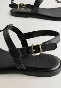 Sandalias de cuero negro con tiras finas, un detallado de hebilla dorada y suela plana. La plantilla está impresa con la marca "NXT".