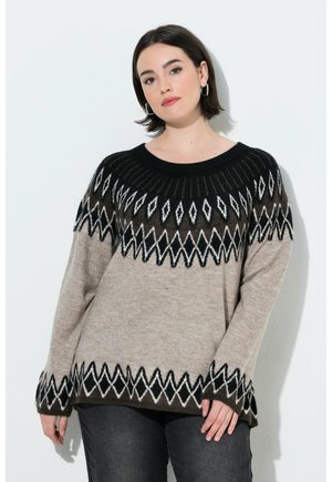Kvinde med kort mørkt hår iført en beige sweater med sorte og hvide geometriske mønstre, der står mod en ensfarvet hvid baggrund.