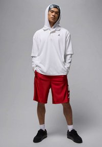 Młody mężczyzna w białej longsleeve polo z kapturem, czerwonych szortach do koszykówki, białych skarpetkach i czarnych tenisówkach, stojący na szarym tle.