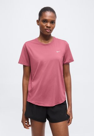 Femme portant un t-shirt rose à manches courtes Nike et un short de sport noir, debout devant un fond blanc uni.