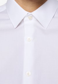 Weißes Hemd mit Knopfleiste, strukturiertem Finish und klassischem Kragen. Es hat sichtbare Knöpfe und ein sauberes, professionelles Design.