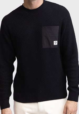 Mann trägt schwarzen strukturierten Strickpullover mit einfarbiger schwarzer Brusttasche und kleinem weißen Logotag, kombiniert mit hellfarbiger Hose.