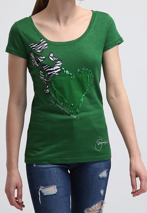 Femme portant un t-shirt vert avec des papillons à rayures zébrées et un cœur en sequins, associé à un jean bleu déchiré.