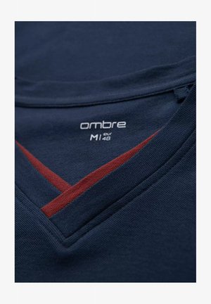 Tessuto blu navy con una finitura strutturata presenta un design a v. Una striscia diagonale rossa evidenzia il colletto. L'etichetta riporta "ombre M1 eur 48."