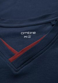 Primo piano di una maglia blu navy con scollo a V, striscia rossa ed etichetta del marchio “ombre” che mostra la taglia M, EUR 48, stampata all'interno.