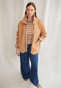 Beige fleece jas met hoge kraag en rits, gelaagd over een beige en witte gestreepte shirt. Losse blauwe denim broek, bruine schoenen.