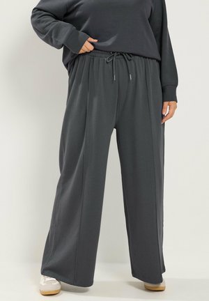 Persona che indossa pantaloni larghi grigio scuro con vita elastica e coulisse, abbinati a scarpe da ginnastica bianche e beige.