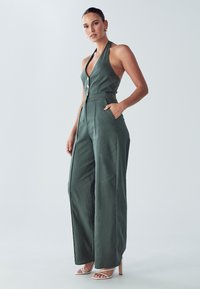 Zelený lněný overal s halter výstřihem, zapínáním na knoflíky vpředu, bočními kapsami, širokými nohavicemi a hladkou texturou. Kombinován s páskovými podpatky.